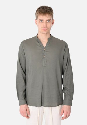 Giovane con capelli corti che indossa una maglia henley a maniche lunghe verde oliva e pantaloni bianchi, in piedi davanti a uno sfondo uniforme.
