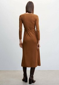 Robe midi à côtes, marron, avec des manches longues et une ceinture à la taille. Elle possède un col arrondi et arrive au mollet, accompagnée de bottes à talons marron.