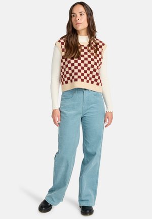 Gehaakte vest met een dambordpatroon in roomkleur en bordeauxrood over een witte lange mouw top. Gekleed met lichtblauwe wijde corduroy broek en zwarte schoenen.