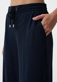 Pantalons de jogger bleu marine avec une taille élastique, fermeture à cordon avec embouts en métal argenté, et poches latérales. Tissu lisse et léger.