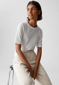 Donna con capelli corti scuri, vestita con una maglietta bianca a maniche corte e pantaloni beige, seduta su uno sgabello nero contro uno sfondo semplice.