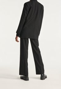 Schwarz gestreifter Anzug mit einem langen Blazer in Slim-Fit und weiten Beinen, aus strukturiertem Stoff. Matte schwarze klobige Stiefel.