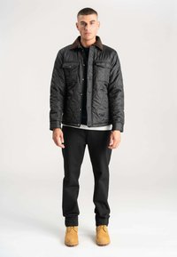 Sort quiltet jakke med brun kordfløjlskrave, to brystlommer foran, båret over en mørk sweater, parret med sorte bukser og brune støvler.