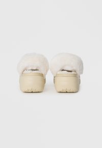 Minkštos off-white šlepetės iš minkštos medžiagos, su apvalia forma ir tekstūruota padanga, užtikrinančia sukibimą. Gale matomas purūs vidus.