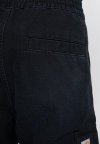Zwarte denim cargobroek met een elastische tailleband, voorzien van een zijzak en een subtiele stoftextuur. Carhartt-logo op de zak.