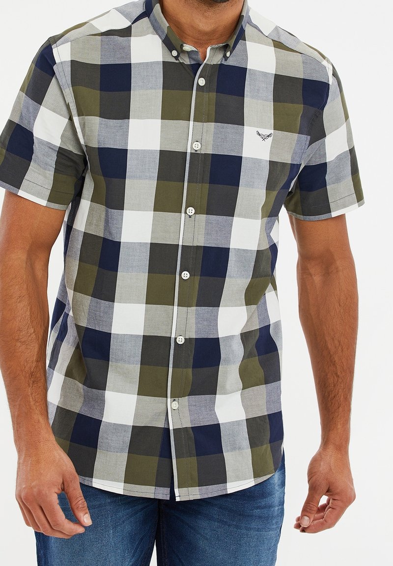 Homme portant une chemise à manches courtes boutonnée avec un grand motif à carreaux en bleu marine, blanc, gris et vert olive, associée à un jean bleu.