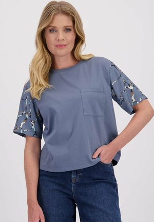 Blonde vrouw in een blauwe shirt met korte mouwen met patroon op de mouwen en een borstzak, gecombineerd met donkerblauwe jeans, met een hand in de zak.