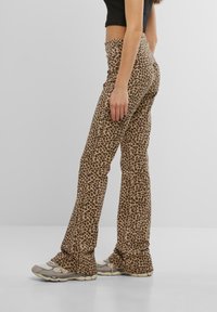Kalhoty s flitry v leopardím vzoru a s vysokým pasem, vyrobené z měkkého materiálu, s béžovým pozadím a tmavě hnědými skvrnami. Rozšířené nohavice sahají až k botám.