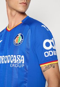Modrá futbalová dres s vertikálnymi pruhmi, červenými a žltými akcentmi na golieri a rukávoch. Obsahuje logo a text na prednej a bočných stranách.