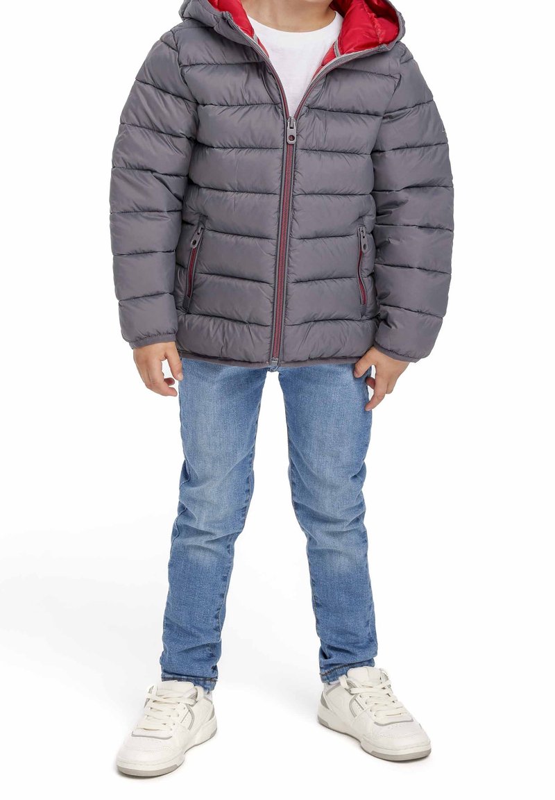 Enfant portant une veste matelassée grise avec une doublure rouge à la capuche, un jean bleu et des baskets blanches, debout sur un fond blanc.