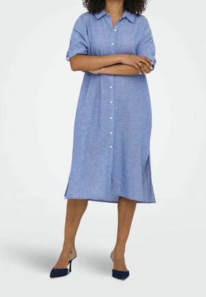 Kvinde iført en blå, stribet knap-ned midi-kjole med op-rullede ærmer og sorte hælede mules, står med armene over kors.