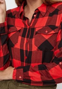 Camicia di flanella a quadri rossi e neri con chiusura a bottoni, due taschini sul petto e bottoni rossi; tessuto leggermente testurizzato.