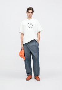 Λευκό κοντο μάνικο μπλουζάκι με γραφικό σχέδιο, φαρδιά παντελόνια τζιν, πορτοκαλί καπέλο κρατημένο στο χέρι και πορτοκαλί δερμάτινα παπούτσια με κορδόνια και χοντρή σόλα.