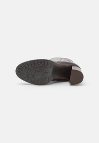 Botte en cuir marron avec une semelle large et texturée présentant un motif de crampons. Le talon est épais et arrondi, offrant une bonne stabilité.