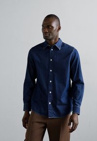 Camicia in denim blu navy con maniche lunghe, colletto button-down e bottoni bianchi; presenta cuciture visibili e un design a vestibilità classica.