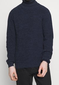 Homme portant un pull à col roulé tricoté texturé bleu marine et un pantalon noir, se tenant devant un fond blanc uni.