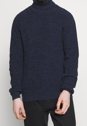 Homme portant un pull à col roulé tricoté texturé bleu marine et un pantalon noir, se tenant devant un fond blanc uni.