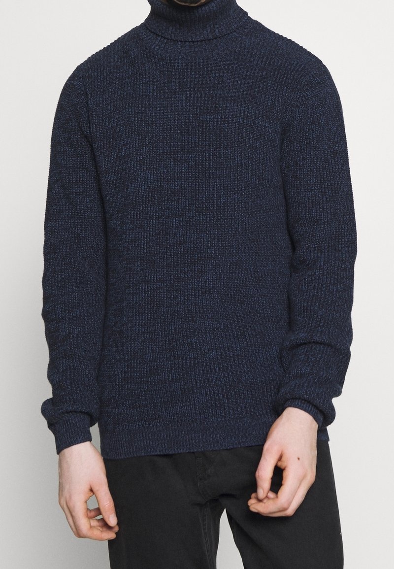 Homme portant un pull à col roulé tricoté texturé bleu marine et un pantalon noir, se tenant devant un fond blanc uni.