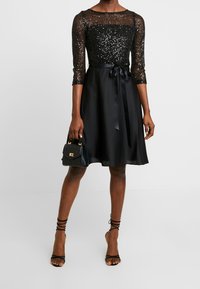 Robe noire avec un corsage à sequins, manches trois-quarts et jupe en satin. Dotée d'une ceinture ruban et accompagnée d'un petit sac à main noir.