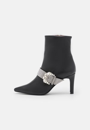 Bottine noire à bout pointu avec talon moyen, ornée d'une bride cloutée de strass et d'une boucle décorative en forme de bijou sur le côté.