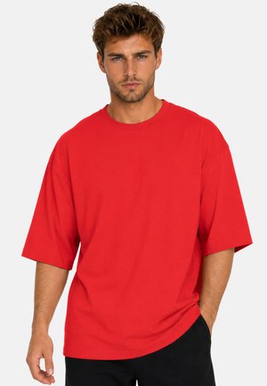 Uomo con capelli corti e ricci che indossa una maglietta rossa larga e pantaloni neri, in piedi davanti a uno sfondo uniforme.