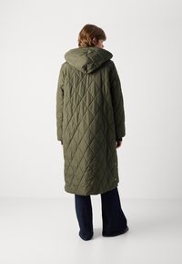 Olijfgroene, gevoerde lange jas met capuchon, voorzien van ruitvormige stiksels en drukknoopaccenten, gecombineerd met donkere flared jeans.