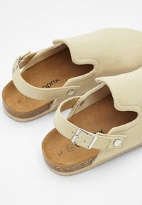 Sandales en daim beige avec un bout arrondi, un dos ouvert et une sangle ajustable. Comprend une semelle en liège et une doublure intérieure lisse. Taille 37.
