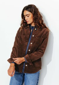 Veste en velours côtelé brun avec deux poches poitrines, devant à boutons et manches retroussées, portée par-dessus une chemise en denim bleu. Tissu texturé.