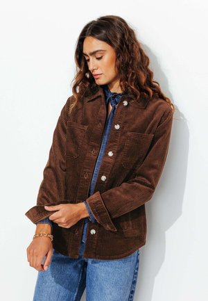 Veste en velours côtelé brun avec deux poches poitrines, devant à boutons et manches retroussées, portée par-dessus une chemise en denim bleu. Tissu texturé.