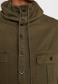 Olivgrön jacka med hög krage, dragsko, framknäppning med knappar och två bröstfickor, med rundade bruna knappar.