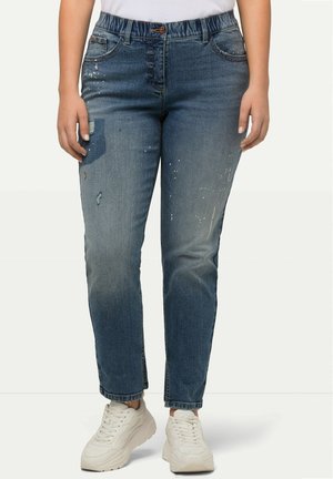 Jeans de mezclilla azul con un corte relajado, que cuentan con una cinturilla elástica, detalles desgastados y un ligero desvanecimiento, combinados con zapatillas blancas.