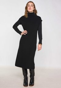 Abito bodycon nero con collo alto, maniche lunghe e silhouette dritta. Texture liscia, abbinato a stivali neri alti fino al ginocchio.