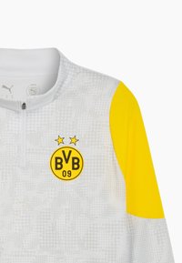 Camiseta atlética blanca de manga larga con un hombro amarillo, con cuello con cremallera. Logo impreso con "BVB 09" y dos estrellas en la parte frontal.