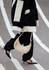 Borsa hobo beige realizzata in pelle liscia, con forma curva, manico corto e dettagli di cucitura discreti. Visibili pantaloni neri e scarpe con tacco.