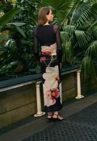 Sort langærmet kjole med blomsterprint, der har et rynket design på ryggen. Tyndhøjede sandaler fuldender outfittet. Grønne planter i baggrunden.