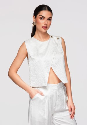 Vrouw die een gestructureerde witte mouwloze top met asymmetrische voorkant draagt en bijpassende hoge taillebroek, poserend met één hand in de zak.