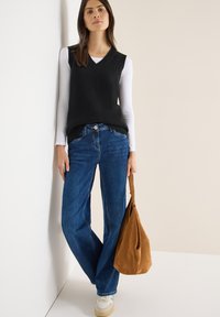 Schwarze Strickweste über einem weißen Langarmshirt, kombiniert mit blauen Jeans mit weitem Bein und einer braunen Wildleder-Hobo-Tasche. Weiße Sneaker vervollständigen das Outfit.