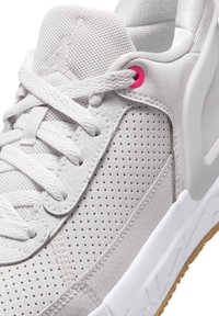 Witte perforatie-sneaker met witte veters, een mesh tong, een roze veteroogje vlak bij de bovenkant en een dikke witte zool met zichtbare bruine buitenzool.