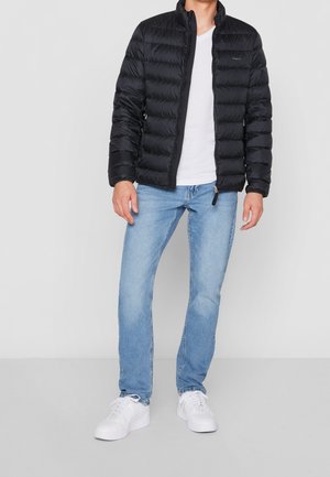 Mężczyzna w czarnej pikowanej kurtce, białej koszulce, jasnoniebieskich dżinsach i białych trampkach stoi na jednolitym tle.