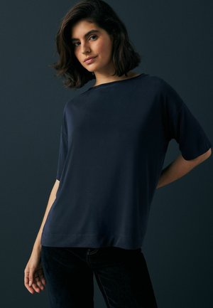 Haut bleu marine à manches courtes avec une coupe détendue et un large décolleté. Fait de tissu lisse, il présente une légère drapé et des détails de couture sur l'ourlet.