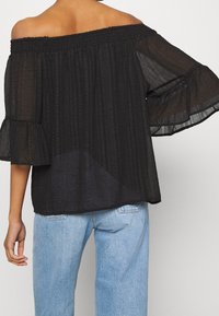Top nero scollato spalle, con tessuto texturizzato, maniche a volant e scollo smock, abbinato a jeans azzurri chiari.