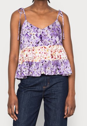 Femme portant un haut sans manches à motifs floraux avec des fleurs violettes et blanches et des bretelles à nouer, associé à un jean bleu foncé.