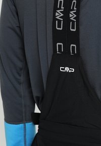 Salopette noire avec bretelles réglables présentant le logo "CMP" en gris ; portée par-dessus un haut à manches longues gris foncé avec des manches à accents bleus.