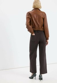 Giacca corta in pelle marrone con orlo e polsini a coste, abbinata a pantaloni a gamba larga marrone scuro e scarpe con tacco verdi. Vista posteriore.