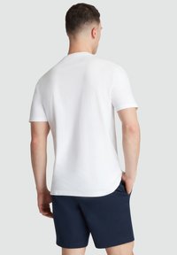 Weißes Kurzarm-T-Shirt mit rundem Halsausschnitt, von hinten präsentiert, kombiniert mit marineblauen Shorts. Glatte Textur, lässiges Design.