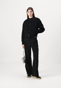 Felpa nera corta con chiusura a bottone, abbinata a pantaloni larghi neri. Le calzature presentano una suola nera e beige, e una piccola borsa metallica.