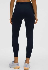Marineblaue, hochtaillierte Leggings aus dehnbarem Stoff mit glatter Textur, ausgestattet mit einer Gesäßtasche und einem Logo am Unterschenkel, kombiniert mit hellen Schuhen.