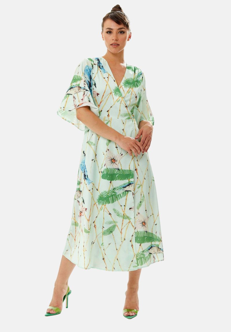 Light green floral wrap dress with short flutter sleeves, featuring a bird and foliage pattern, paired with green high-heeled sandals.