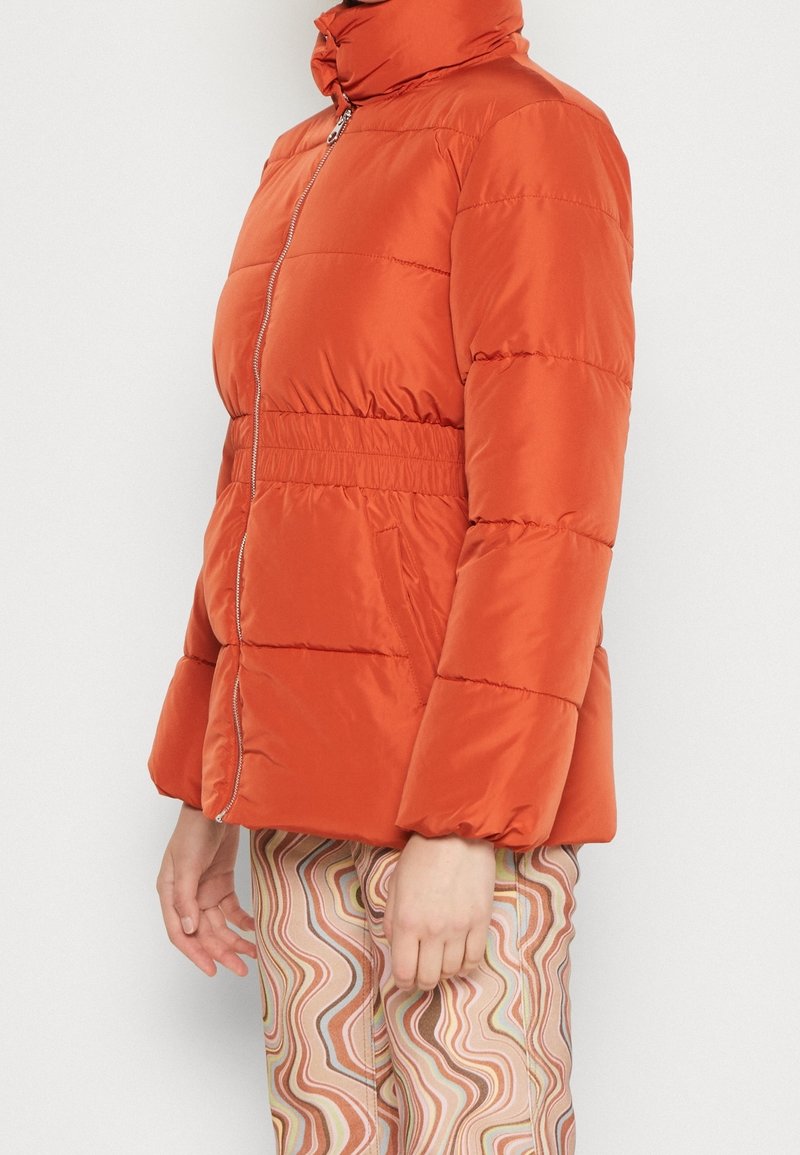 Personne portant une doudoune orange vif avec fermeture éclair devant et taille élastique, associée à un pantalon taille haute avec des motifs ondulés multicolores.