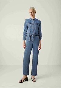 Denimoverall i ljusblått, med insvängd midja, lång ärm, knappfront och rak ben-design. Svarta klacksandaler fulländar looken.